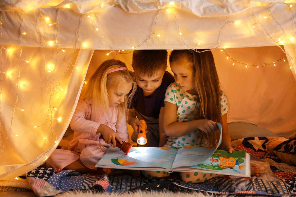 2 fetite 1 baiat citesc carte cu lanterne in cort acasa
